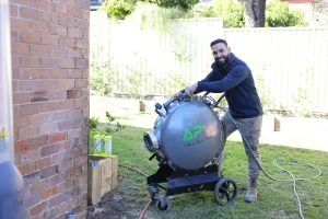 A man in a garden operates a large, cylindrical metal tank on wheels labeled "AP" beside a brick wall.