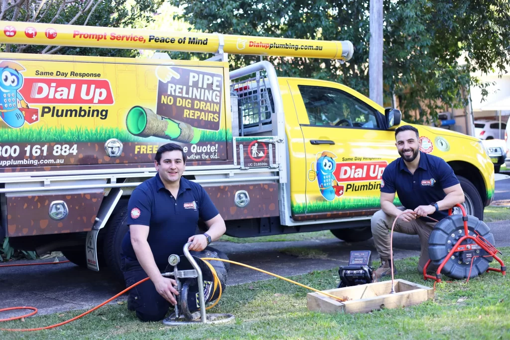 Two plumbers kneeling near plumbing equipment in front of a Dial Up Plumbing service truck, offering pipe relining and repairs.