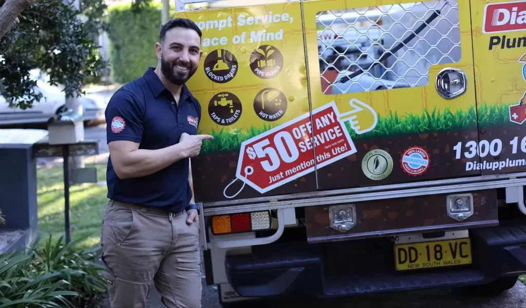 Plumber pointing to a promotional sign on a utility vehicle offering $50 off any service. The truck advertises plumbing services.