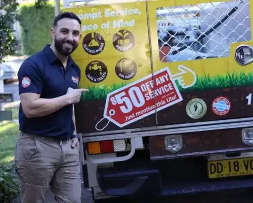 Man in blue shirt and khaki pants points to a "50% off any service" ad on the back of a parked truck with various service icons.