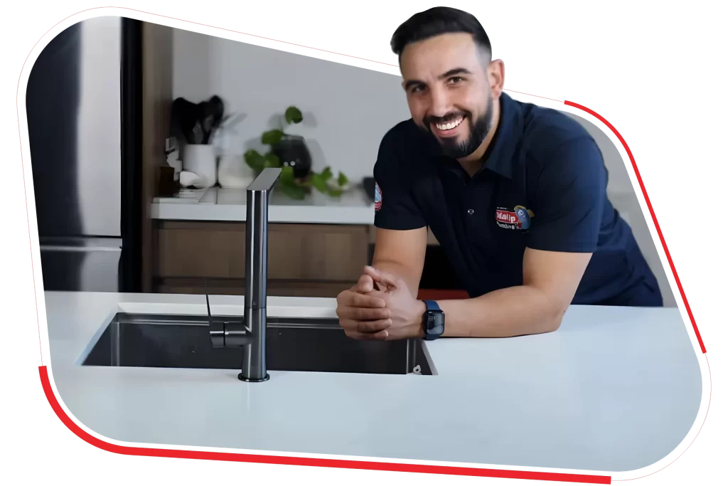 Smiling man in a logoed shirt leans on a modern kitchen counter near a faucet, with plants and kitchenware in the background.