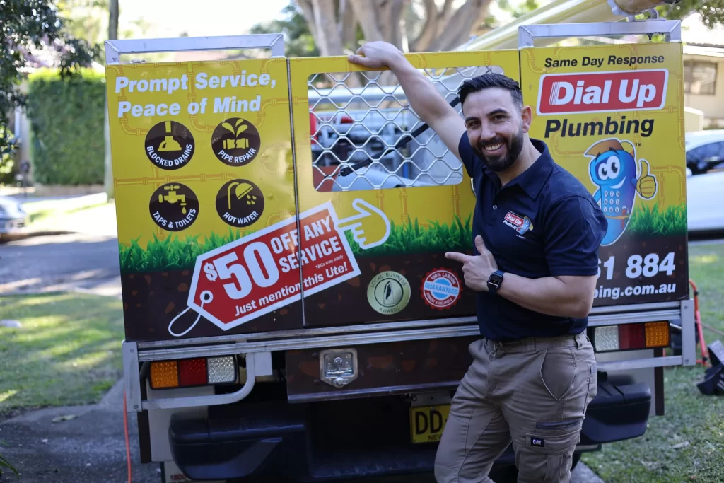 Smiling plumber stands next to a company vehicle displaying Dial Up Plumbing service details and a $50 off service promotion.