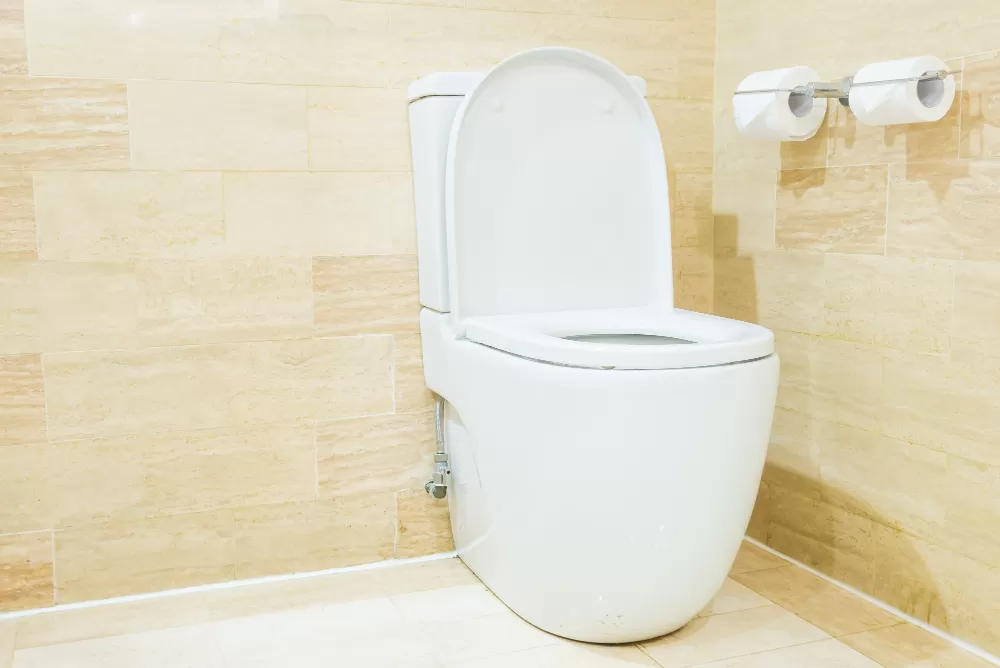 Modern white toilet with an open lid in a beige tiled bathroom, accompanied by two toilet paper rolls on a holder.